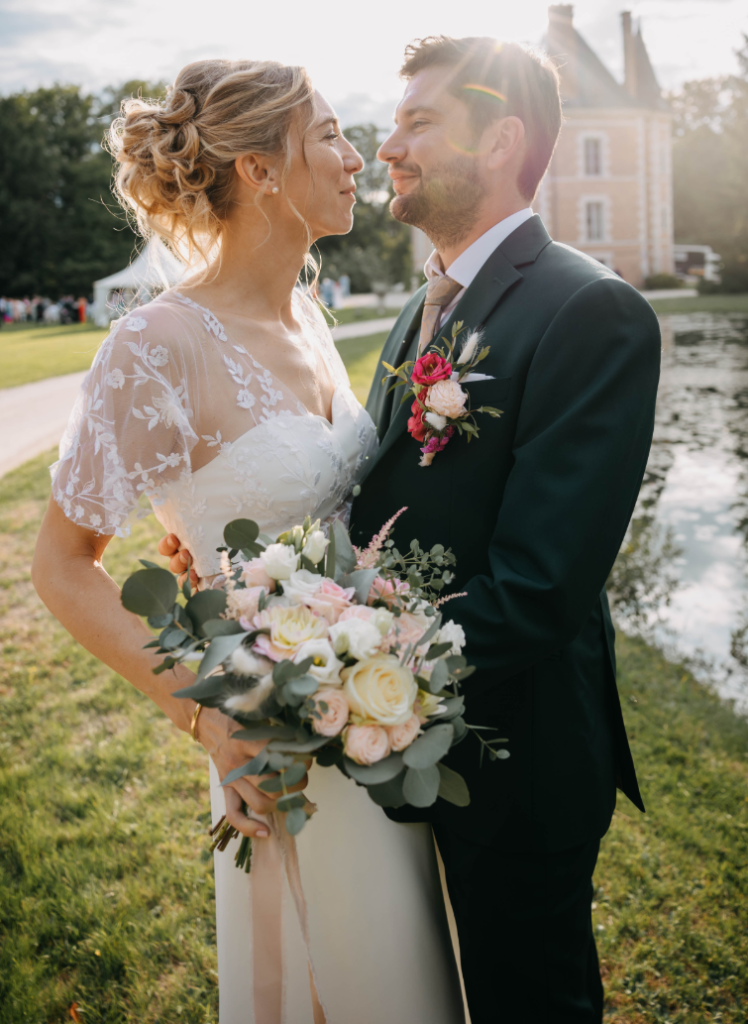 Couple de mariés en extérieur : marié en costume vert sur mesure avec boutonnière florale, posé près d’un plan d’eau.