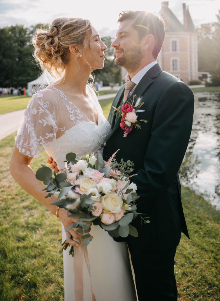 Couple de mariés en extérieur : marié en costume vert sur mesure avec boutonnière florale, posé près d’un plan d’eau.